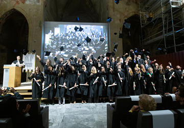 Graduates in black gowns celebrate by throwing their caps in the air during a commencement ceremony, with a large screen displaying the audience behind them.